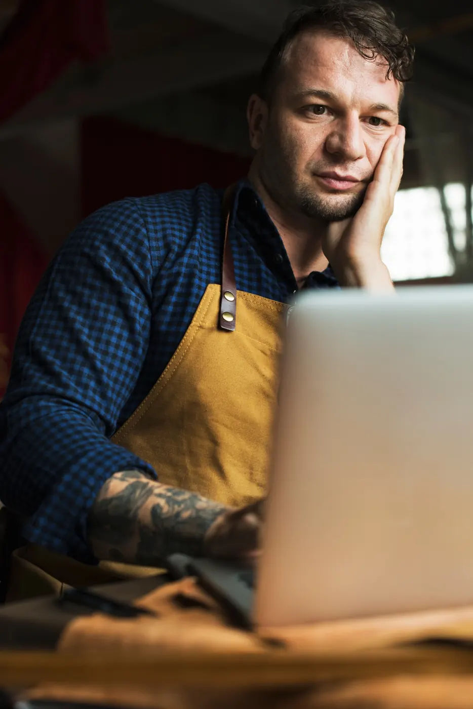 A shop owner in an apron thinking in front of his laptop