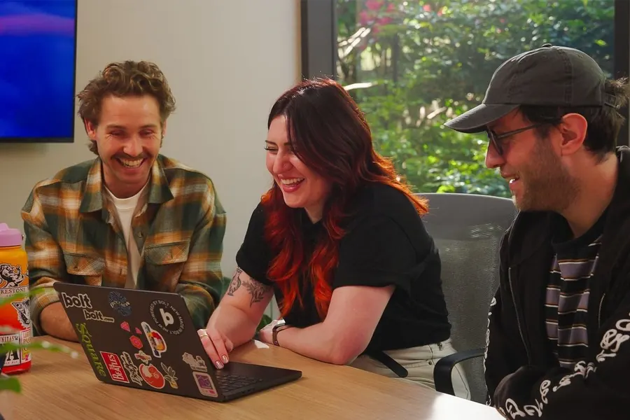Three teammates laughing around a laptop in a meeting room